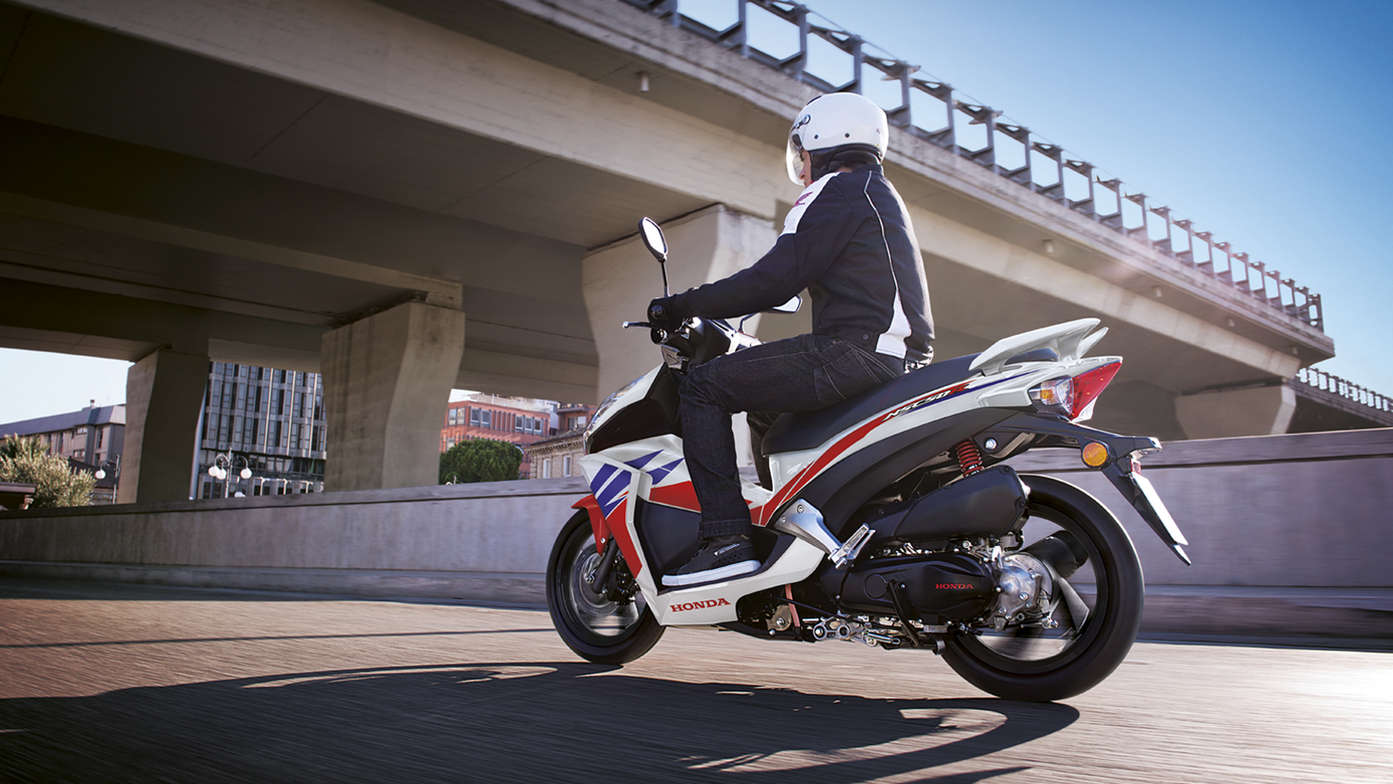 Repsol Honda Vision scooter rear-threequarter view with rider. Left-facing (Road location). 