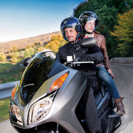 Front 3-quarter view of Forza 300 with rider and pillion, road location.